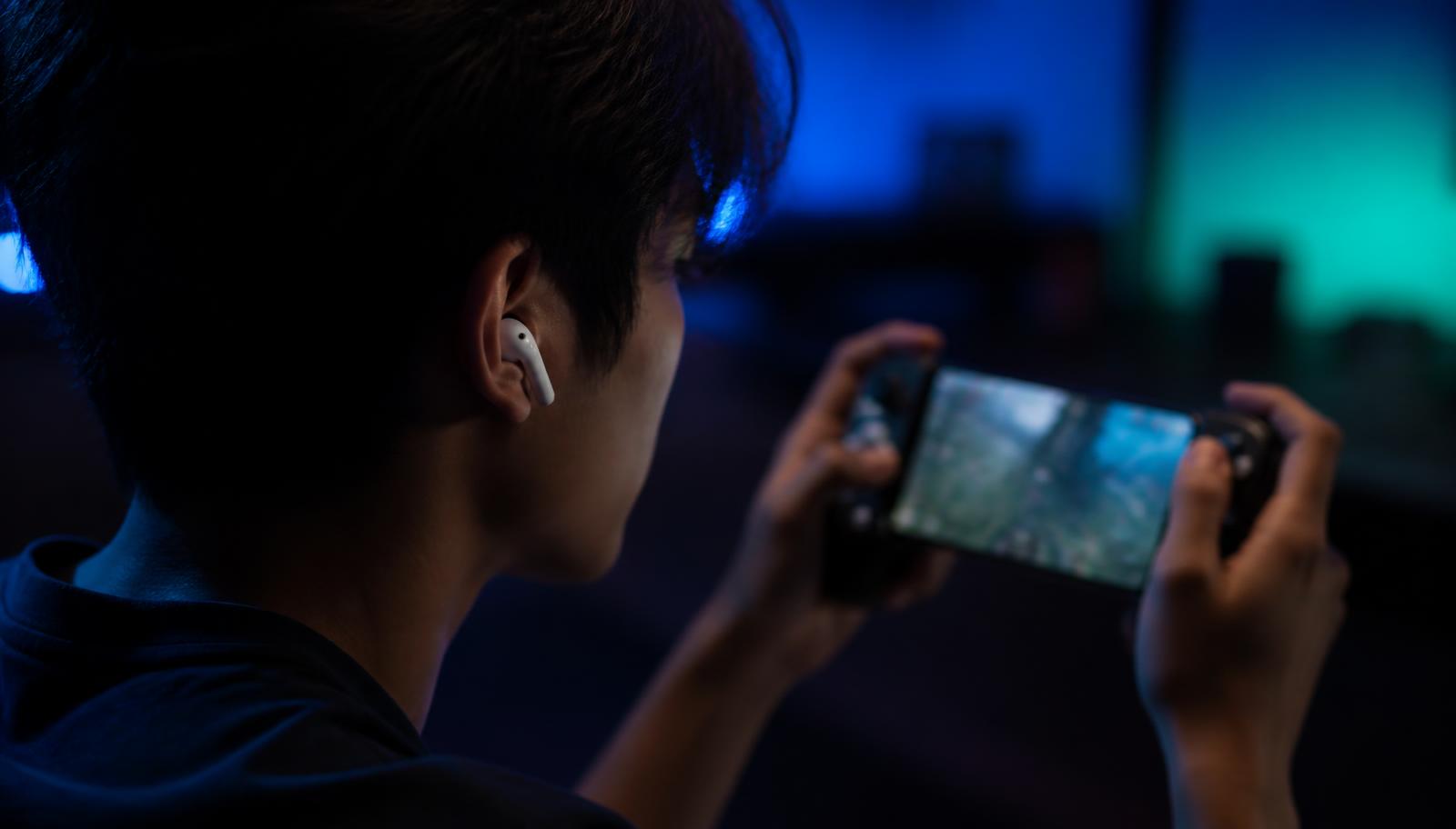 Over-the-shoulder view of a player wearing white wireless earbuds and holding a phone in a controller grip in a blue-lit room