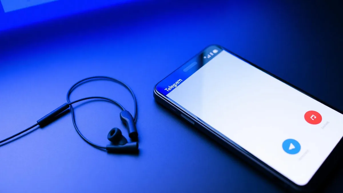A smartphone showing an active Telegram voice call screen next to a pair of wired in-ear earbuds on a dark blue surface