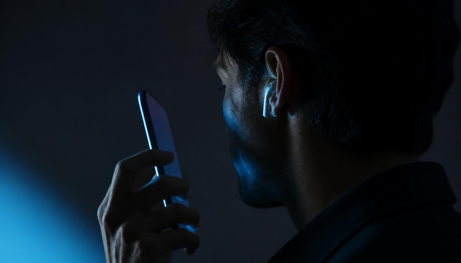 A person on a phone call in low blue light, wearing a wireless earbud, phone screen turned away from the camera